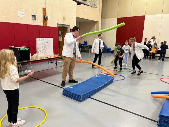 Physician assistant majors from WSU play with kids from Paper Mill Elementary School. One student in a white lab-coat uses foam noodles to simulate an obstacle course while a kid tries to jump over it.
