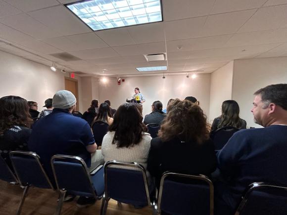 A shot of a student reading poetry at the Chapbook Presentation event on December 13. Rows of people sit in front in blue chairs, with the speaker reciting the poetry into a microphone.