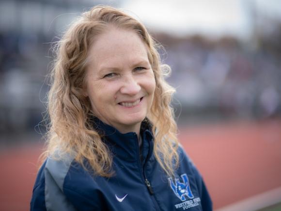 Nancy Bals, Associate Director of Athletics, smiles and poses on the University's track, which is blurred out. She is wearing a long-sleeved, navy sweater with the University's Owl logo on it.