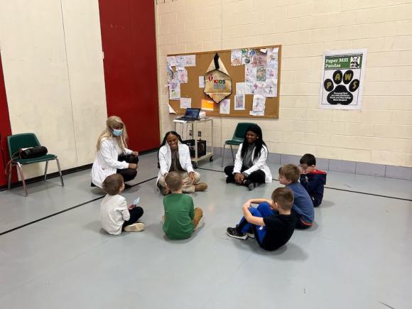 Three physician assistant majors from WSU in white lab-coats sit in a circle with children from the Paper Mill Elementary School in Westfield as part of an all-day presentation, where the PA students teach kids about germs and wellness.
