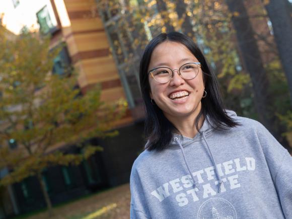 Student wearing grey WSU sweatshirt with white writing wearing glasses smiling with fall trees in background.