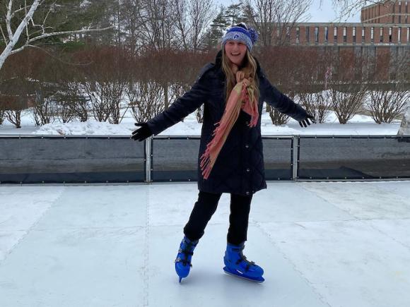 The faux ice-skating rink available for students during the weekend festivities. A woman student in a black coat, peach scarf, blue hat, and navy blue skates glides on top of the fake ice.