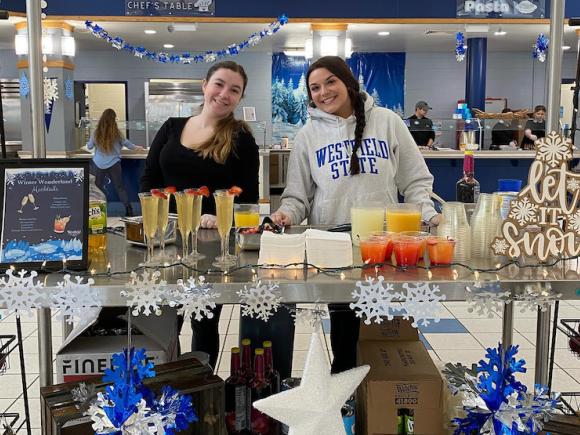 Two young women serve colorful cocktails in the Dining Commons. The table they're at are decorate with blue and white snowflakes, and they are standing in the middle of the Dining Commons.