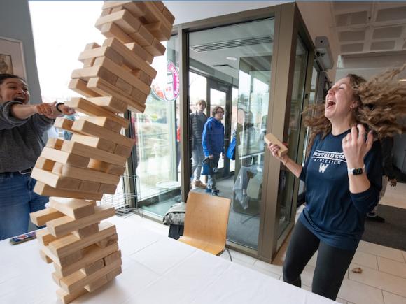 Two students in the bottom level of Ely play jenga, with wooden blocks. The tower is mid-fall, with the two women laughing. Both wear dark blue T-shirts and dark pants.