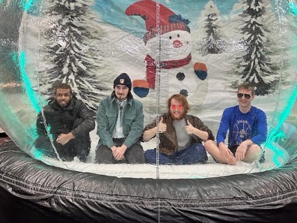 Four students, all young men, sit inside a giant, inflatable snow globe as part of the campus' Winter Weekend. One student wears sunglasses while another wears a hat, and they're all smiling in front of the snowman decoration inside the globe.