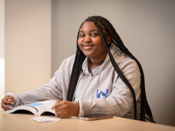 Jakayla Leary, a first-year at the University, sits at a desk with a book open before her. Her long brown hair is down, and she wears a grey sweatshirt with a blue Nestor logo on the front.