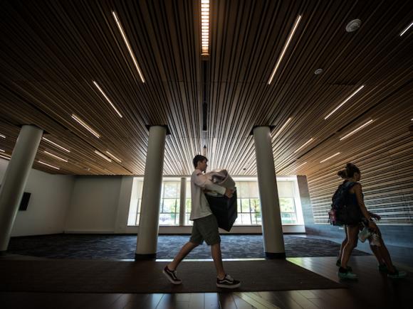 A shot of the lobby of New Hall. Two students are carrying backpacks and walking through it. The room is lowly lit, with the overhead lights photographed with a wide lens.
