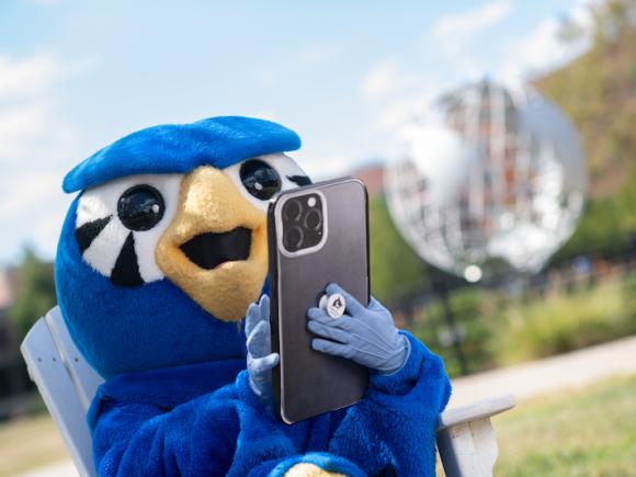 Nestor, the school mascot, wears his blue Owl suit. Nestor is holding a large phone in his hands and sits in front of the campus globe.