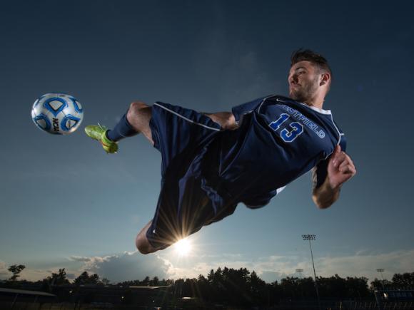 A close-up shot of a student from 2014 in a blue, Owl, soccer team uniform. He is mid-air, leg extended and about to kick a ball that was passed to him.