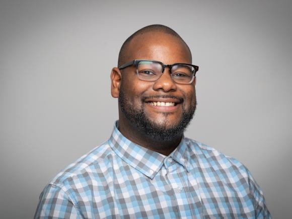 Joseph Seal, Academic Support Coordinator for the Course Achievement, Retention, and Engagement Center. He wears a white and blue-checkered button-down shirt and black glasses. He smiles in front of a plain, gray background.