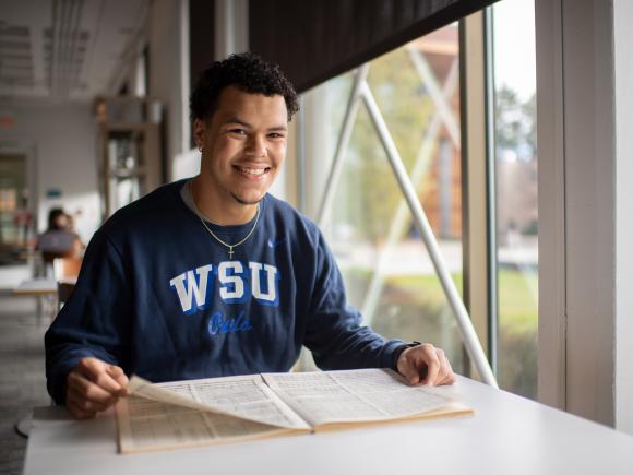 TRIO student wearing blue WSU shirt smiling with book open.