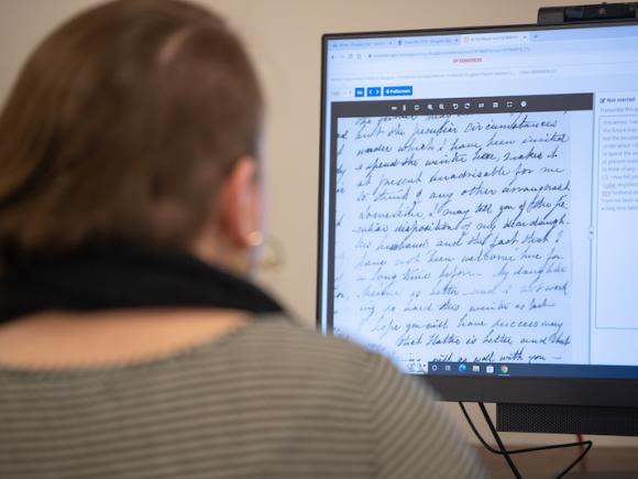 A close-up of a student participating in Douglass Day, 2024. Their back is turned to the camera and is blurry, though they are wearing a gray sweater and dark blue scarf. They sit at a desktop computer, which shows historical documents from the 19th century as part of Douglass Day.