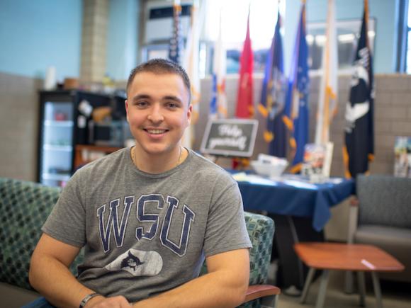 Ethan Lichwan, class of 2025. He is in the Veteran and Military office on campus, and wears a gray shirt with a blue "WSU" print on the front. He has close-cropped hair. Colored flags are blurred in the background and he is sitting on a green couch.