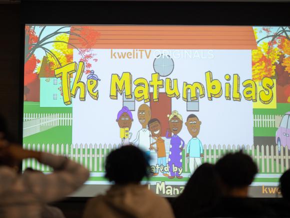 A close-up of a projector screen. "The Matimbilas", an animation about a Tanzanian-American family, is on the screen. The title is in a blocky, yellow font, and the animated family pose in front of a white house. Several silhouettes can be seen in front of the projector.
