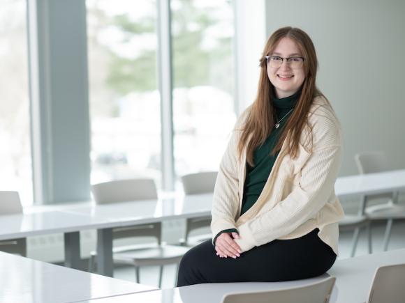 Nicole Dolat, class of 2024. She is dressed in a green shirt, a white over-sweater, black glasses, black pants, and sits on top of a white, glossy desk. The walls behind her are white, and she is in a classroom. Windows along it are open, and there is greenery blurred in the background.