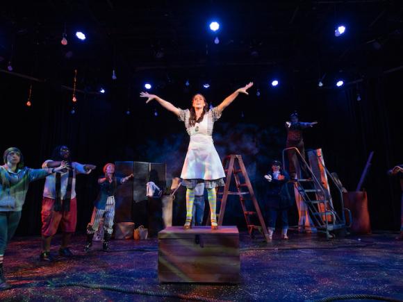 Westfield State's production of "Peter and the Starcatcher". A lone student wearing a blue dress is standing on top of a wooden chest on stage. Their hands and arms are splayed out above their head, at their sides. Several other actors surround them as they perform.