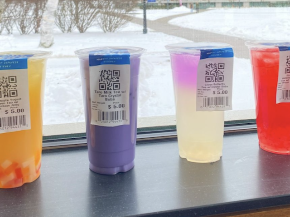 Four boba tea, orange, purple, clear, and red colored, rest on a black windowsill which overlooks the campus green. There's fresh snow on the ground and shrubbery in the background next to the stone walkway.