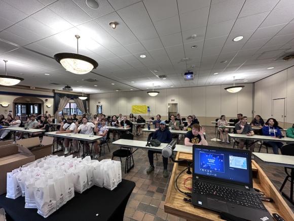 As part of RA Appreciation Week, a room full of Res sit at thin tables while being recognized for their efforts. A table with a black tablecloth holds prizes in white bags for each RA to celebrate them. An open laptop rests on a podium at the front of the room.