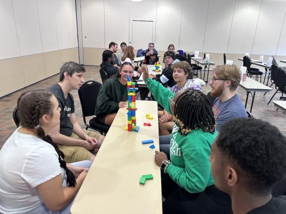 For RA Appreciation Week. Several students sit at a long, thin table, where they're playing jenga, a block-building game. They're being honored as part of RA Appreciation Week.