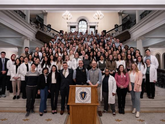 2024 Lobby Day, an event for Physician Assistants and students studying to be PAs. A large group of people, some in white coats, gather on the steps of the venue in Boston, MA. A wooden podium sits before all of them. Two chandeliers hang in the background behind them.