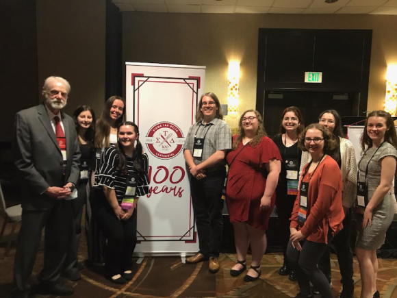 The 2023 Sigma Tau Delta Centennial Convention. Dr. Glenn Brewster, along with Alexis Crafts, Sam Grunden, Chloe Sanfacon, Victoria Nesmelova, and Julia Robak. They stand, pose, and smile in front of a small white banner which says, "100 years" in red script. 