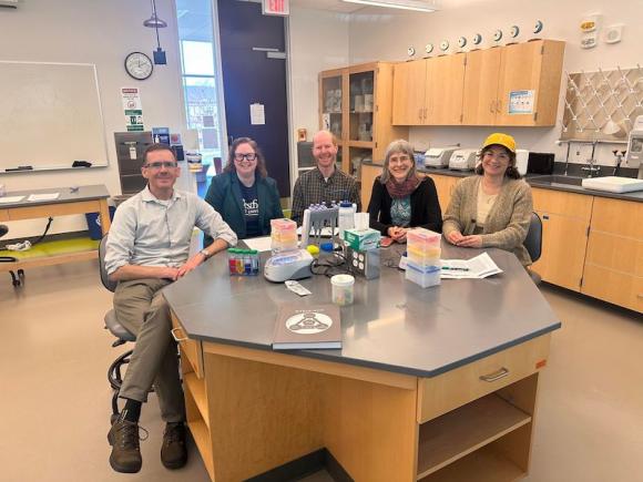 From left to right: Dr. Arne Christensen, Dr. Megan Kennedy, Dr. Volker Ecke, Dr. Christine Von Renesse, and Dr. Shannon Gleason. They sit in a Biology lab, surrounded by tan desks and cabinets along the walls. They sit together at one desk, wearing blue lanyards and smiling at the camera as part of an interview on the University's new STEM-ACT grant.