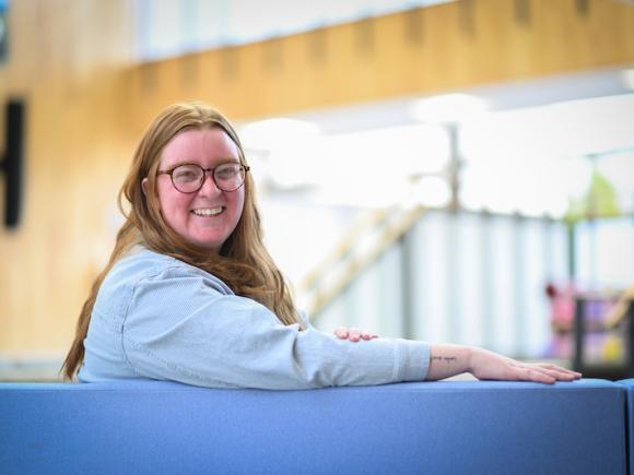 Averie Bye-Dickinson, a graduate student of the Master go Public Administration program. She is sitting in the lobby in Horace Mann, and is wearing a gray, long-sleeve shirt. She has red hair and glasses, and has one arm over the back of the chair she sits on as she smiles at the camera. White light from the background streams in behind her.