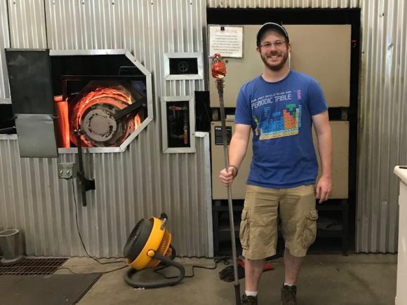 Brian Mernoff, an alum of the University. He is standing next to heavy machinery and is wearing a blue T-shirt and tan cargo shorts. He's smiling while holding a staff.