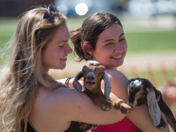 Fresh Check Day 2023. Two women students hold baby goats as part of learning about self-care. One has long, blond hair while the other has brown hair in a ponytail. The goats have brown fur and are nestled in their arms.