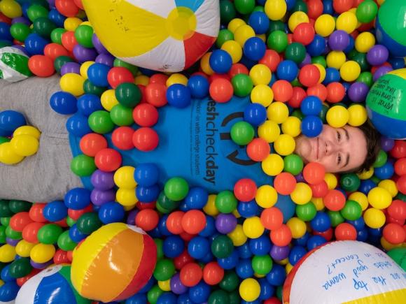 A male student from the 2023 Fresh Check Day. He is wearing a dark blue shirt with black lettering that says, "Fresh Check Day", and is laying in a colorful ball pit while smiling at the camera.