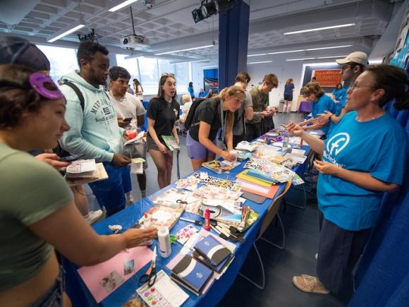 Fresh Check Day 2023. A crowd of students stand in front of a booth promoting mental health wellness. A professional wearing a blue T-shirt which says, "Fresh Check Day" is speaking to them. Paper and various arts and crafts are on the surface of the booth.