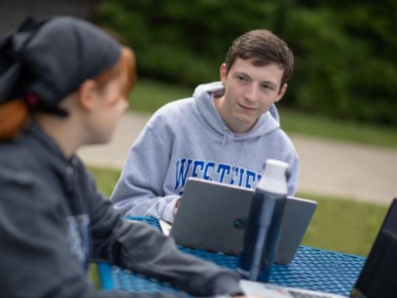 Two students work on their laptops while seated at a table outside on the Westfield State University campus.