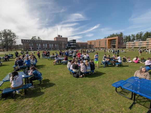 The 2024 total solar eclipse. The photo is of the campus green before the Ely building. Clusters of students sit together across the green in preparation for the eclipse. They sit either on the ground or on blue tables dotting throughout the field.