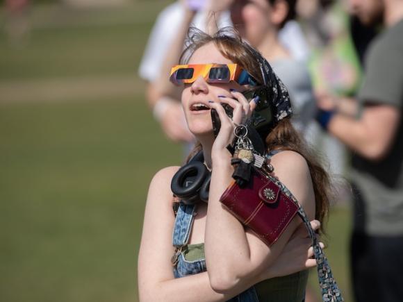 A student looking at the 2024 total solar eclipse. She has her phone to her ear and eclipse glasses on her face. Her mouth is open in awe as she looks at the sky.