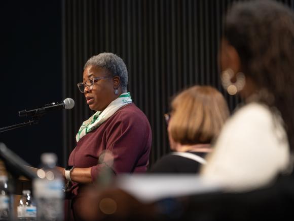 Kimberly Williams, from the the Center for Human Development. She is wearing a maroon, long-sleeved jacket with a white scarf around her neck. She is on stage, and speaking into a microphone as part of the "Supporting Our Superwomen" symposium.