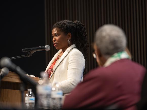 Cheryl Woods Giscombe, a professor at The University of North Carolina at Chapel Hill. She is on stage standing into a microphone. She wears a white, long-sleeved jacket and a pink undershirt. Her hair is pulled back into a ponytail.