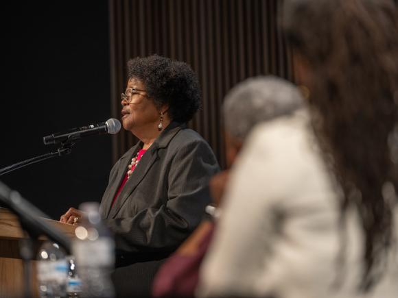 Phyllis Sharps, from the Johns Hopkins University School of Nursing. She is wearing a black, long-sleeved jacket with a red undershirt. She is on stage, and speaking into a microphone as part of the "Supporting Our Superwomen" symposium.