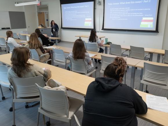 From the 2024 WMWP's Best Practices for Teachers conference. A woman presents her research before two rows of participants. Two projector screens are on the wall as she presents.