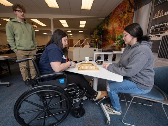 Occupational therapy students. Two sit across from each other on a white table and play jingo. Another stands by and watches.