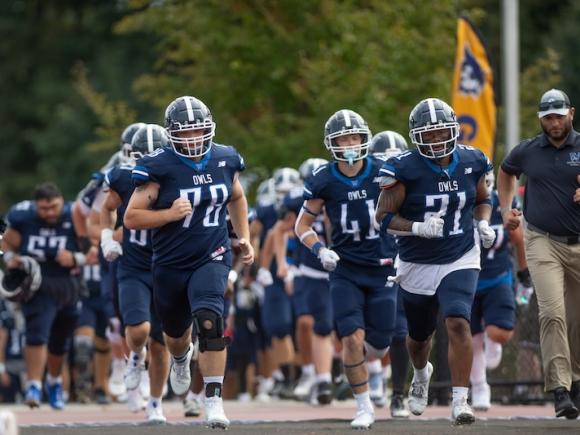 The Westfield State Owl Football team on the 2024 Family Weekend football game. Lines of students in their dark blue uniforms jog out after being introduced.