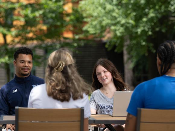 Four college students sitting outdoors with laptops, studying on a sunny day.
