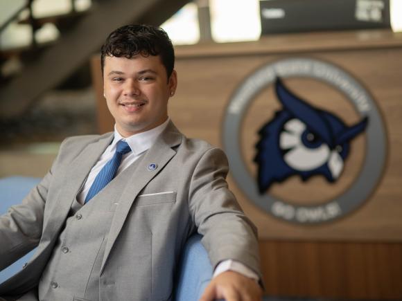 Westfield State student in a gray suit smiles while seated in front of a university podium featuring the Westfield State owl logo.
