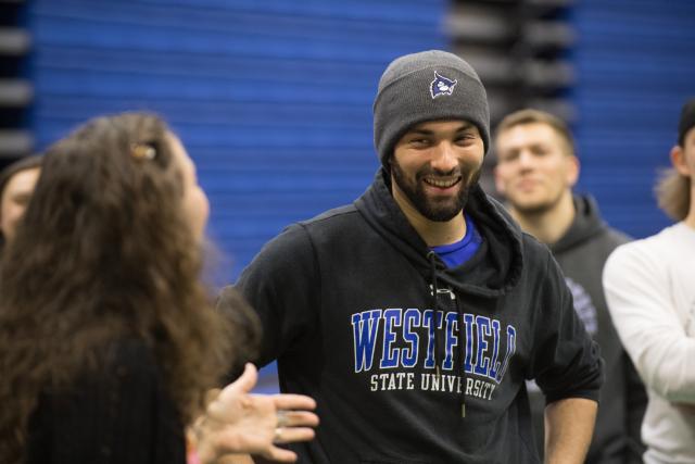 Male student in WSU beanie and sweatshirt, smiles as his classmate gives a in-class presentation