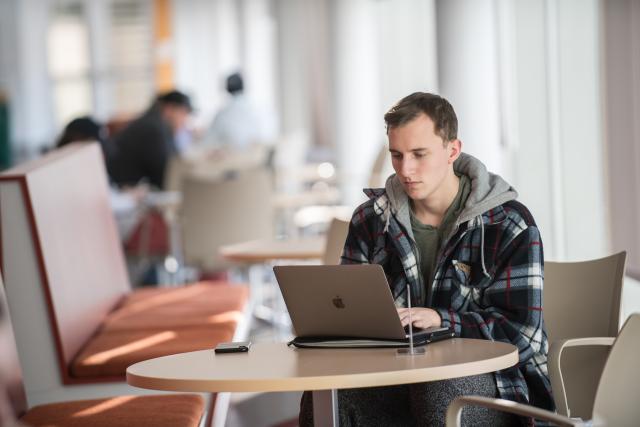 Male student works on his laptop at a table inside the Stevens Center