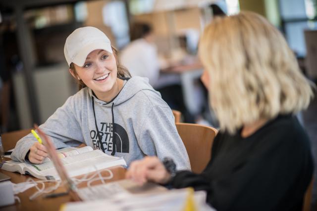 Female student in white ball cap smiling at friend as they work in the Ely library