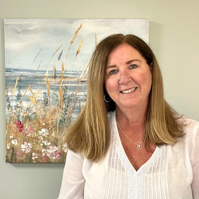 A white woman is standing in front of a beach painting and smiling