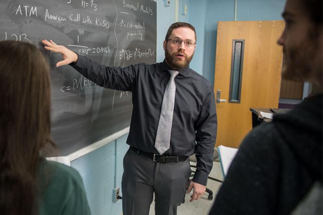 A finance instructor motions to notes written on a chalkboard while talking to two students.