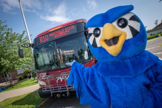 A photo of Nestor in his blue, owl costume and pointing towards a red bus which has the university's address and route on the front.