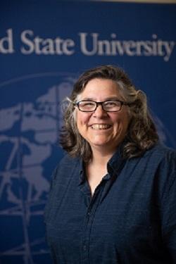 Elisabeth Stassinos, Department Chair Headshot Wearing Glasses with Westfield State screen in the back.
