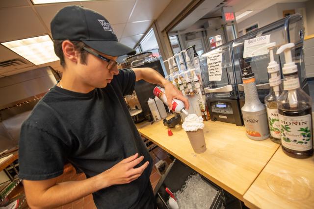 Owl cafe staff member wearing black WSU hat with logo putting whip cream on top of iced coffee.